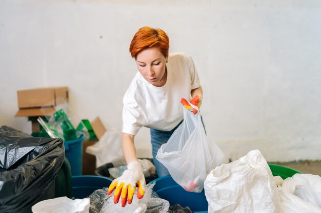 Ernsthafte, hübsche, rothaarige Arbeiterin mit Latexhandschuhen, die verschiedene Abfälle zur weiteren Entsorgung in einer modernen privaten Abfallrecyclinganlage sortiert