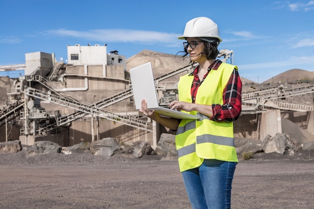 Ernsthafte Ingenieurin im Helm und in der Weste, die Informationen auf Laptop suchen, während sie in der Nähe der Industrieanlage der Baustelle stehen