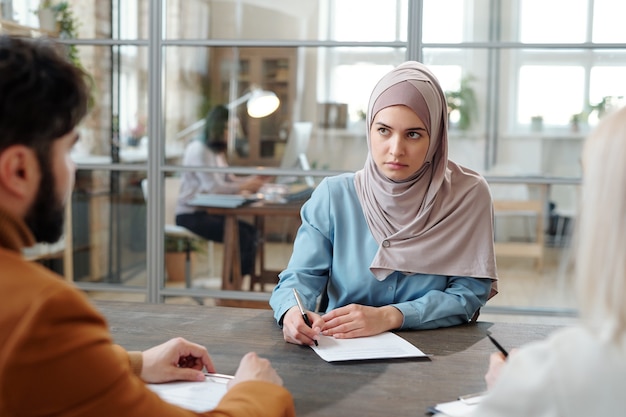 Ernsthafte junge arabische Frau im Hijab, die vor Personalmanagern am Tisch sitzt und beim Vorstellungsgespräch einen Fragebogen ausfüllt