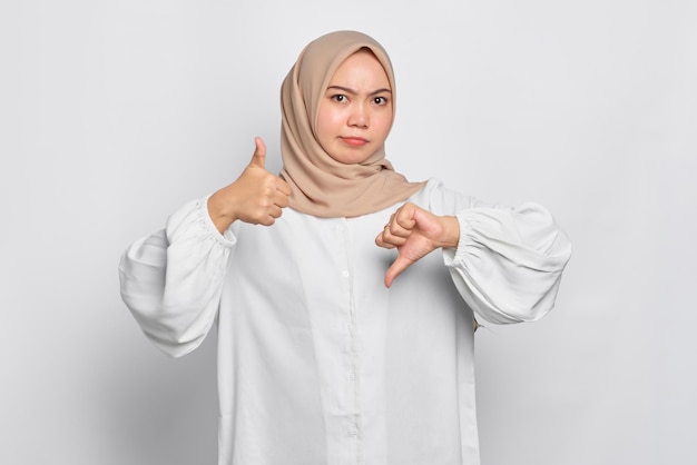Ernsthafte junge asiatische muslimische Frau zeigt Daumen nach oben und unten Entscheidung isoliert auf weißem Hintergrund