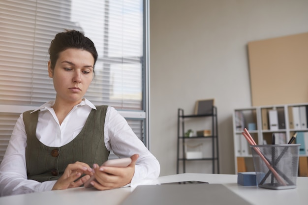 Ernsthafte junge Frau, die am Tisch im Büro sitzt, schreibt eine Nachricht mit ihrem Handy