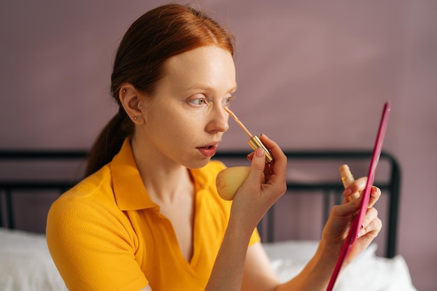 Ernsthafte junge Frau, die Concealer auf das Gesicht unter den Augenringen aufträgt, um die Haut zu korrigieren, sitzt im Schlafzimmer auf dem Bett. Porträt einer rothaarigen Frau, die Make-up macht und Grundtoncreme mit Foundation aufträgt