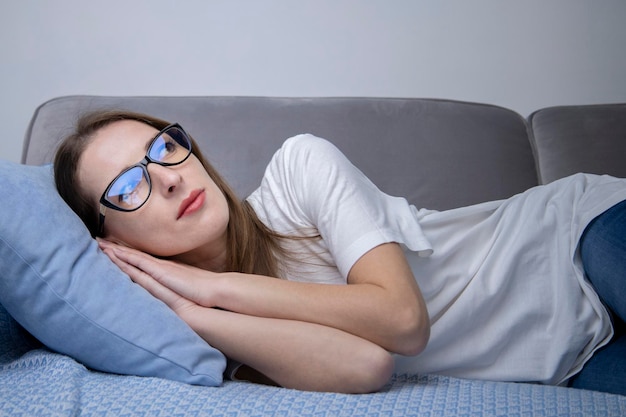 Ernsthafte junge Frau im weißen T-Shirt mit Brille auf dem Sofa liegend