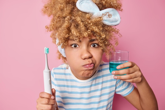 Foto ernsthafte junge frau mit buschigen haaren in einem lässig gestreiften t-shirt hält eine elektrische bürste und ein glas frisches mundwasser kümmert sich um die mundhygiene, die über rosafarbenem hintergrund isoliert ist mundhygiene