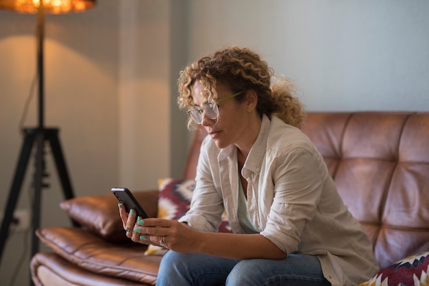 Ernsthafte junge Frau SMS mit Handy beim Sitzen auf dem Sofa zu Hause. Frau mit lockigem Haar und Brille liest oder schreibt mit dem Handy im Wohnzimmer