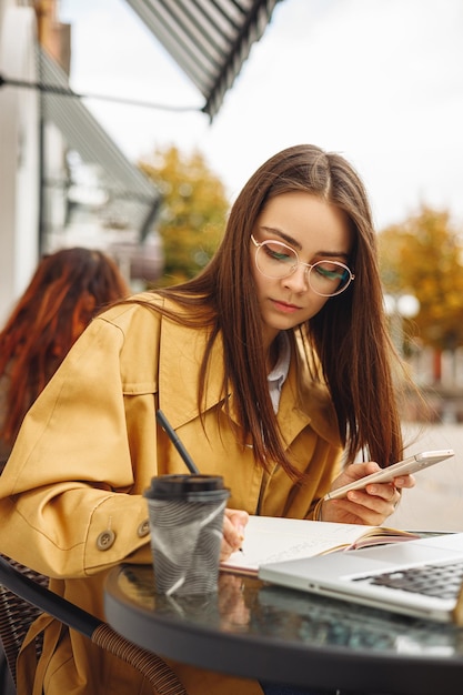 Ernsthafte junge Freiberuflerin, die in Notizbuch schreibt und Handy benutzt, während sie am Tisch arbeitet