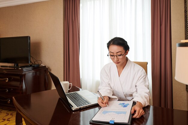 Ernsthafte junge Geschäftsfrau im Bademantel, die den Verkaufsbericht analysiert, wenn sie am Tisch im Hotelzimmer sitzt