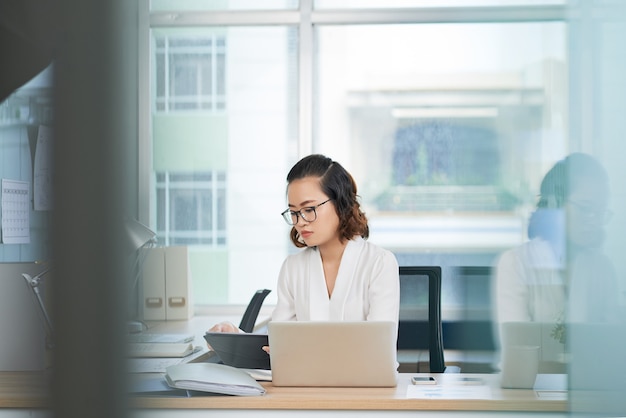 Ernsthafte junge Geschäftsfrau mit Brille, die am Bürotisch sitzt und einen Bericht oder einen Artikel über den Talet-Computer liest