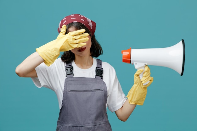 Ernsthafte junge Putzfrau mit einheitlichem Bandana und Gummihandschuhen, die den Lautsprecher hält, der den Lautsprecher zur Seite zeigt und die Hand vor den Augen isoliert auf blauem Hintergrund hält