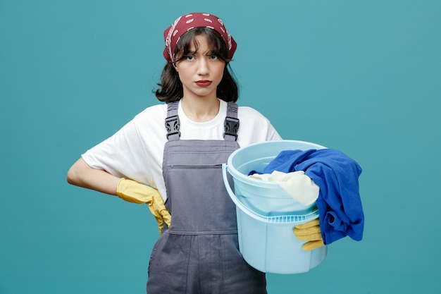 Ernsthafte junge Putzfrau mit einheitlichem Kopftuch und Gummihandschuhen, die einen Eimer mit schmutziger Wäsche hält und in die Kamera schaut, während sie die Hand auf der Taille isoliert auf blauem Hintergrund hält