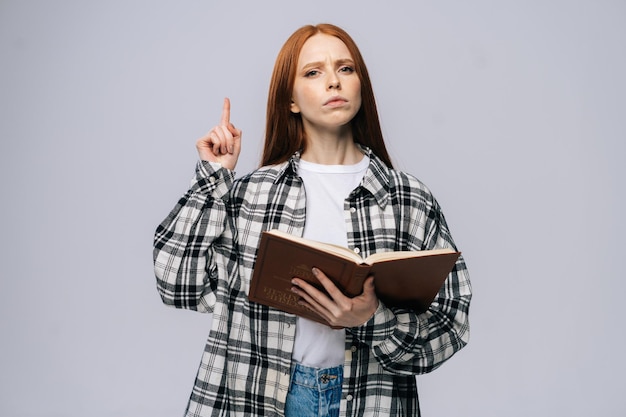 Ernsthafte junge Studentin, die ein Buch hält und mit dem Finger nach oben zeigt und auf grauem, isoliertem Hintergrund in die Kamera schaut. Hübsche rothaarige Dame, die legere Kleidung trägt und emotional Gesichtsausdrücke zeigt