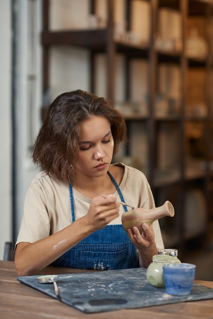 Ernsthafte junge Töpferin mit Schmutz im Gesicht, die am Schreibtisch sitzt und Tontopf in der Werkstatt malt