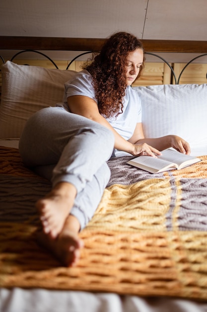 Ernsthafte kaukasische, lockige, brünette Frau, die auf einem mit Frottee-Tagesdecke bedeckten Bett ruht, mit konzentriertem Gesicht, das ein Buch in den Händen hält und tagsüber einen Schlafanzug trägt. Beruhigende Freizeitaktivität