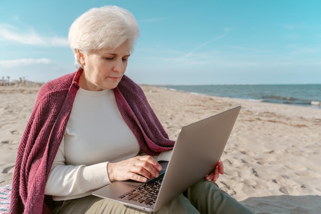 Ernsthafte konzentrierte attraktive kaukasische grauhaarige Dame, die am Strand sitzt und auf dem Laptop tippt