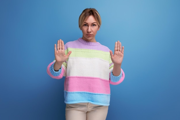 Ernsthafte konzentrierte blonde junge Frau, die Stoppschild mit der Hand auf blauem Hintergrund zeigt