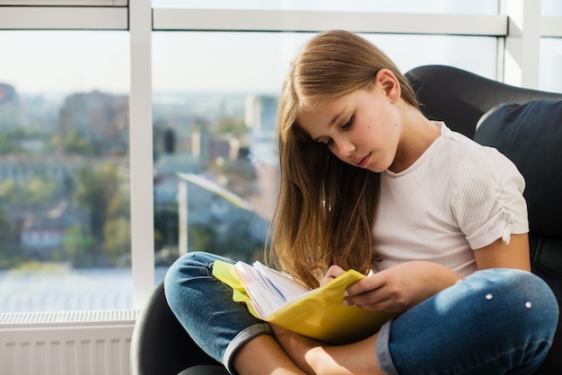 Ernsthafte konzentrierte jugendliche Studentin, die das Buch zu Hause liest, mit großen Fenstern im Sonnenlicht auf einem bequemen schwarzen Stuhl in Lotus-Pose mit lässigem Jeans-Outfit barfuß
