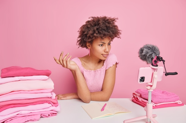 Ernsthafte lockige Afro-Amerikanerin sieht Tutorial-Video über Smartphone an, macht sich Notizen, wie man Wäsche wäscht, schreibt die Waschtemperatur verschiedener Kleidungsstücke auf, sitzt am Tisch gegen die rosa Wand
