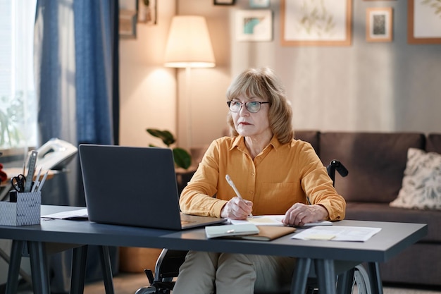 Ernsthafte reife Frau mit Brille, die am Tisch sitzt und auf den Bildschirm des Laptops schaut, sie arbeitet online und macht sich Notizen im Dokument, sie arbeitet zu Hause