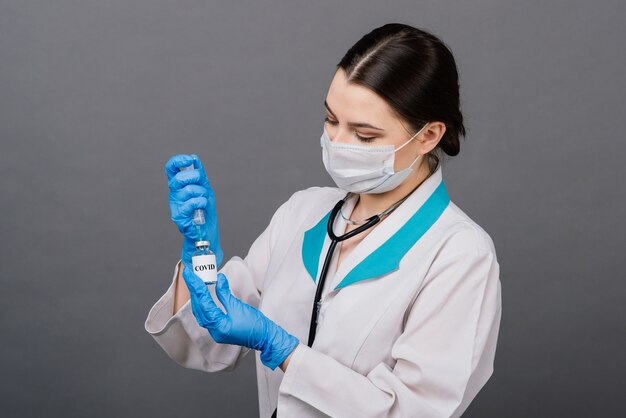 Ernsthafte Ärztin in einer medizinischen Maske mit Spritze und Impfstoff isoliert auf Grau