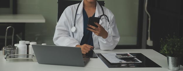 Ernsthafte Ärztin mit Laptop und Notizen in der medizinischen Zeitschrift sitzt am Schreibtisch Junge Ärztin in weißem Mantel und Stethoskop arbeitet am Computer am Arbeitsplatz