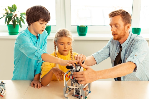 Ernsthafte Schulkinder, die mit ihrem Lehrer für Naturwissenschaften am Schreibtisch sitzen und mit ihrem Lehrer einen Roboter aus Bausatz anschalten