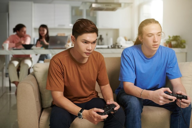 Ernsthafte Teenager spielen zu Hause Videospiele, während ihre Freunde im Hintergrund Hausaufgaben machen