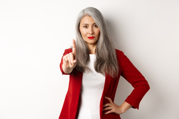 Ernsthafte und enttäuschte asiatische Geschäftsfrau, die eine Stopp-Geste zeigt, den Finger missbilligt, etwas Schlechtes ablehnt, auf weißem Hintergrund steht