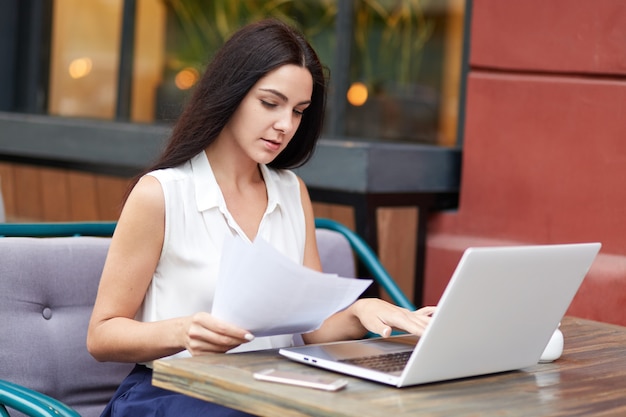 Ernsthafte Unternehmerin konzentriert sich auf Dokumente, liest aufmerksam die Vertragsbedingungen, benutzt einen Laptop und verbringt die Essenszeit im Straßencafé.