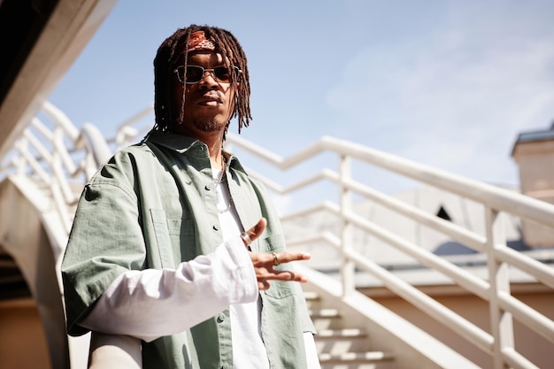 Ernsthafter afroamerikanischer Typ mit Sonnenbrille und stilvoller Freizeitkleidung steht gegen Geländer moderner Architektur und blauen Himmels in einer städtischen Umgebung