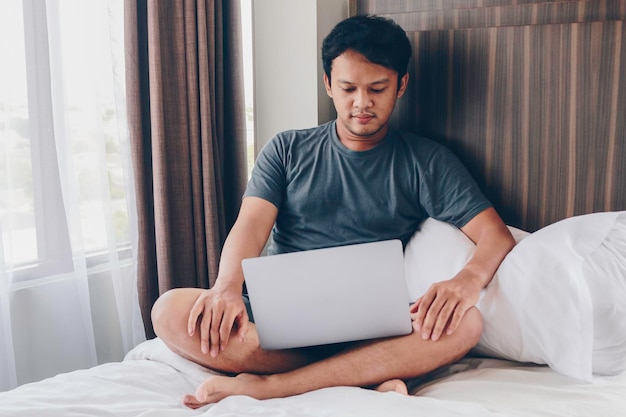 Ernsthafter asiatischer Mann arbeitet mit seinem Laptop auf seinem Bett Konzept eines erfolgreichen Lebensstils für Freiberufler
