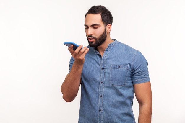 Ernsthafter bärtiger Mann in blauem Hemd, der das Handy in der Hand hält und dem Sprachassistenten Befehle gibt, die eine Sprachnachricht hinterlassen oder Audionotizen aufnehmen Studioaufnahme im Innenbereich isoliert auf weißem Hintergrund