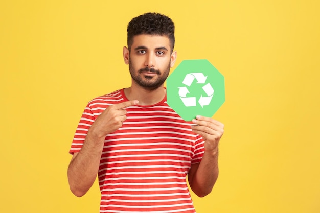 Ernsthafter bärtiger Mann in rotem T-Shirt, der mit dem Finger auf das Abfallrecycling-Symbol in seiner Hand zeigt und sich Sorgen um Ökologie und Umweltverschmutzung macht. Studioaufnahme im Innenbereich isoliert auf gelbem Hintergrund