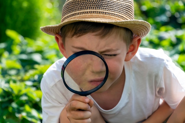 Ernsthafter entzückender kleiner Kinderjunge in Strohhut mit Lupe, der beobachtet oder sucht. Kind führt Ermittlungen durch, unterzieht sich einer Suche. Kleiner Detektiv.