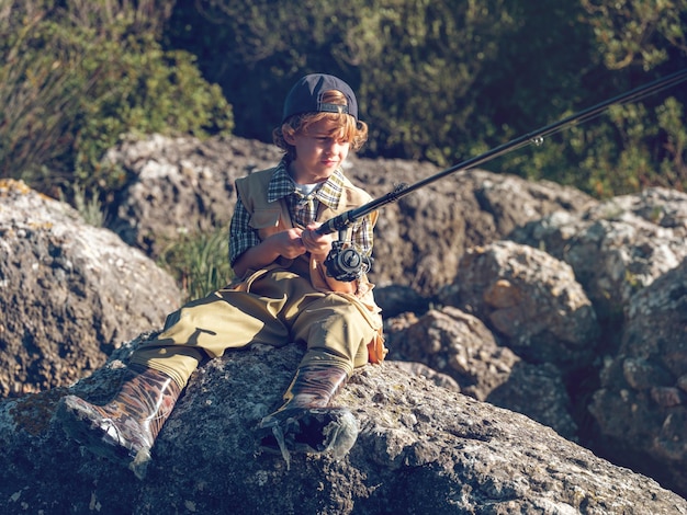 Ernsthafter Ganzkörperjunge in lässiger Kleidung und Mütze, der wegschaut und fischt, während er an einem sonnigen Tag auf dem Land am steinigen Ufer sitzt