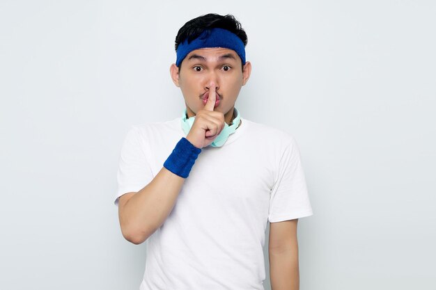 Ernsthafter junger asiatischer Sportler mit blauem Stirnband und weißem Sportbekleidungs-T-Shirt mit Kopfhörern, die ein Zeichen der Stillegeste zeigen, die Finger in den Mund steckt, isoliert auf weißem Hintergrund