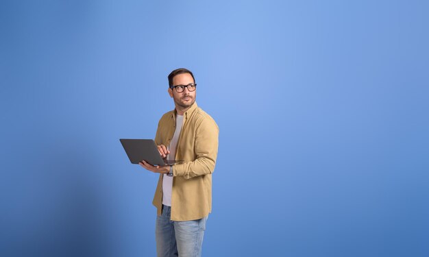 Ernsthafter junger Geschäftsmann hält einen Laptop in der Hand und blickt nachdenklich über einen abgeschiedenen blauen Hintergrund weg