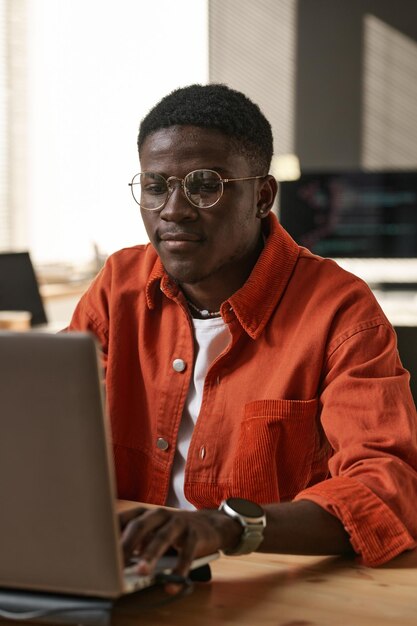 Ernsthafter junger IT-Support-Ingenieur in Freizeitkleidung und Brille sitzt am Arbeitsplatz vor dem Laptop