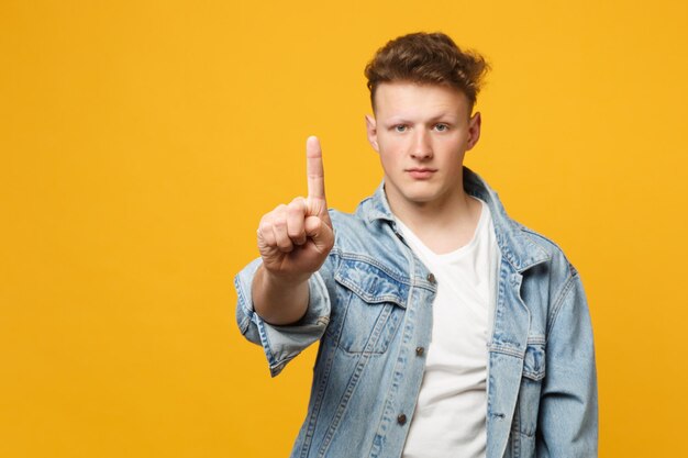 Ernsthafter junger Mann berührt so etwas wie einen Klick auf den Knopf und zeigt auf einen schwebenden virtuellen Bildschirm, der auf gelb-orangefarbenem Hintergrund isoliert ist. Menschen aufrichtige Emotionen, Lifestyle-Konzept. Mock-up-Kopienbereich.