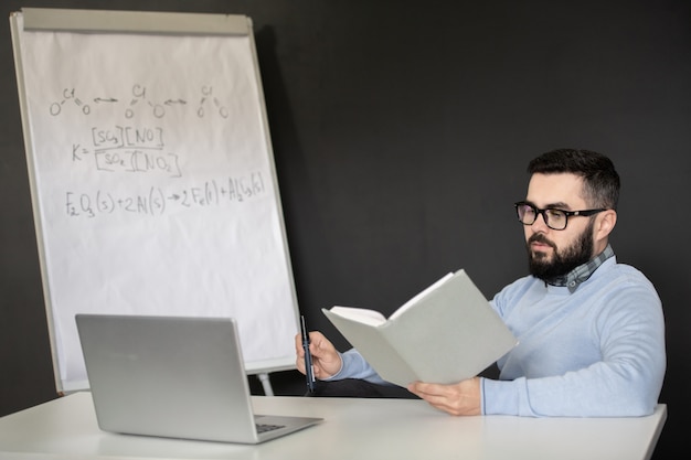 Ernsthafter junger Mann im Lesebuch der Brille und der Freizeitkleidung beim Sitzen vor Laptop während der Online-Lektion der Chemie zu Hause