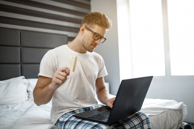 Ernsthafter junger Mann sitzt heute Morgen auf dem Bett. Guy trägt Pyjama und Brille. Er hält die Kreditkarte in der Hand und tippt auf der Laptoptastatur.