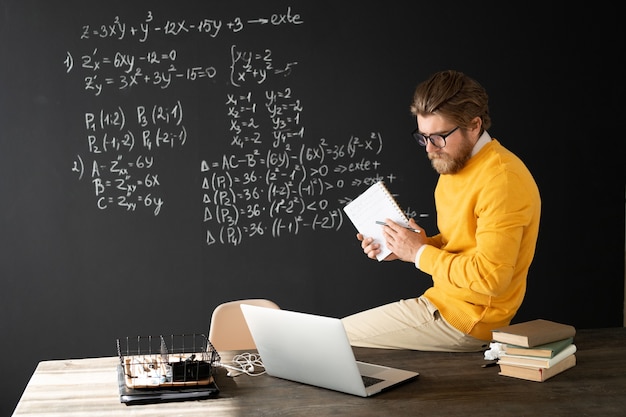 Ernsthafter Lehrer mit Heft, das auf Gleichung auf Tafel mit Kreide zeigt, während er Online-Schülern erklärt, wie man es löst