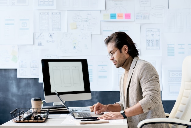 Ernsthafter Manager der App-Designfirma, die am Tisch im Büro mit Plantafel sitzt und Laptop für Arbeit verwendet