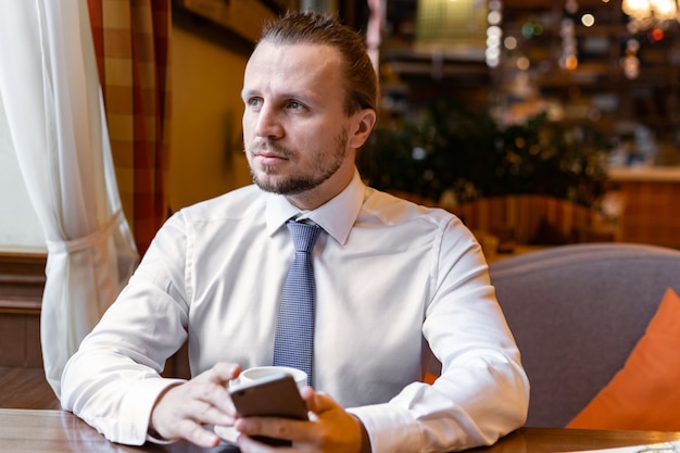 Ernsthafter Mann, der in der Kaffeestange sitzt und ein Handy während der Kaffeepause in der Hand hält.