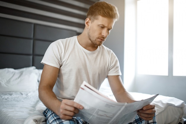 Ernsthafter nachdenklicher junger Mann am Bett am frühen Morgen. Er las Zeitung.