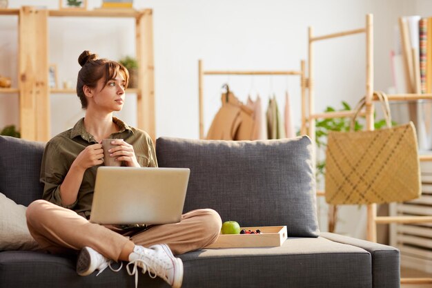 Ernsthafter, nachdenklicher, kreativer Designer mit Haarknoten, der mit gekreuzten Beinen auf dem Sofa im Heimbüro sitzt und