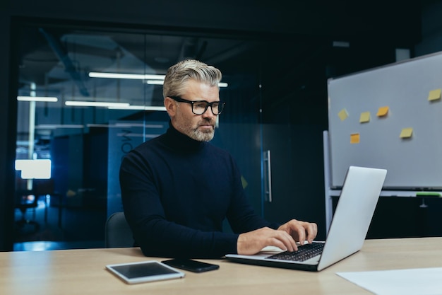 Ernsthafter, reifer, grauhaariger Geschäftsmann, der in einem modernen Büro arbeitet und nachdenklich auf dem Laptop tippt