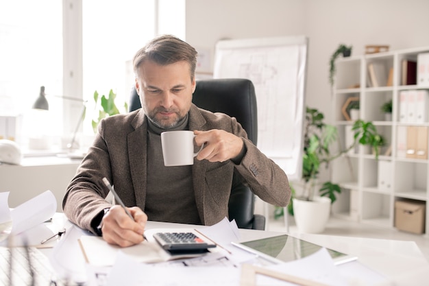 Ernsthafter reifer Ingenieur oder Buchhalter mit Stift und Tasse Kaffee, die Berechnungen und Notizen im Notizbuch im Büro machen