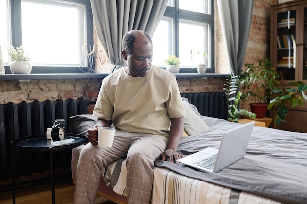 Ernsthafter schwarzer Mann im Ruhestand mit einer Tasse Kaffee, der die Online-Nachrichtenrolle durchsieht