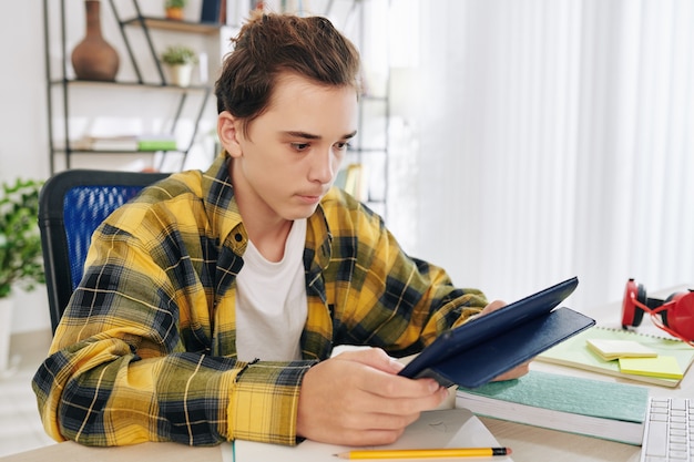 Ernsthafter Teenager, der Studentenbuch auf Tablet-Computer liest und Homeschooling wegen Coronavirus-Pandemie