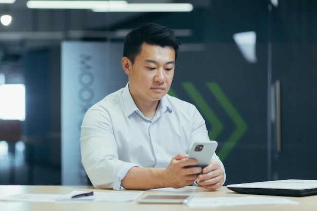 Ernsthafter und nachdenklicher asiatischer Geschäftsmann mit Telefon, der in einem modernen Büro arbeitet und am Telefon tippt